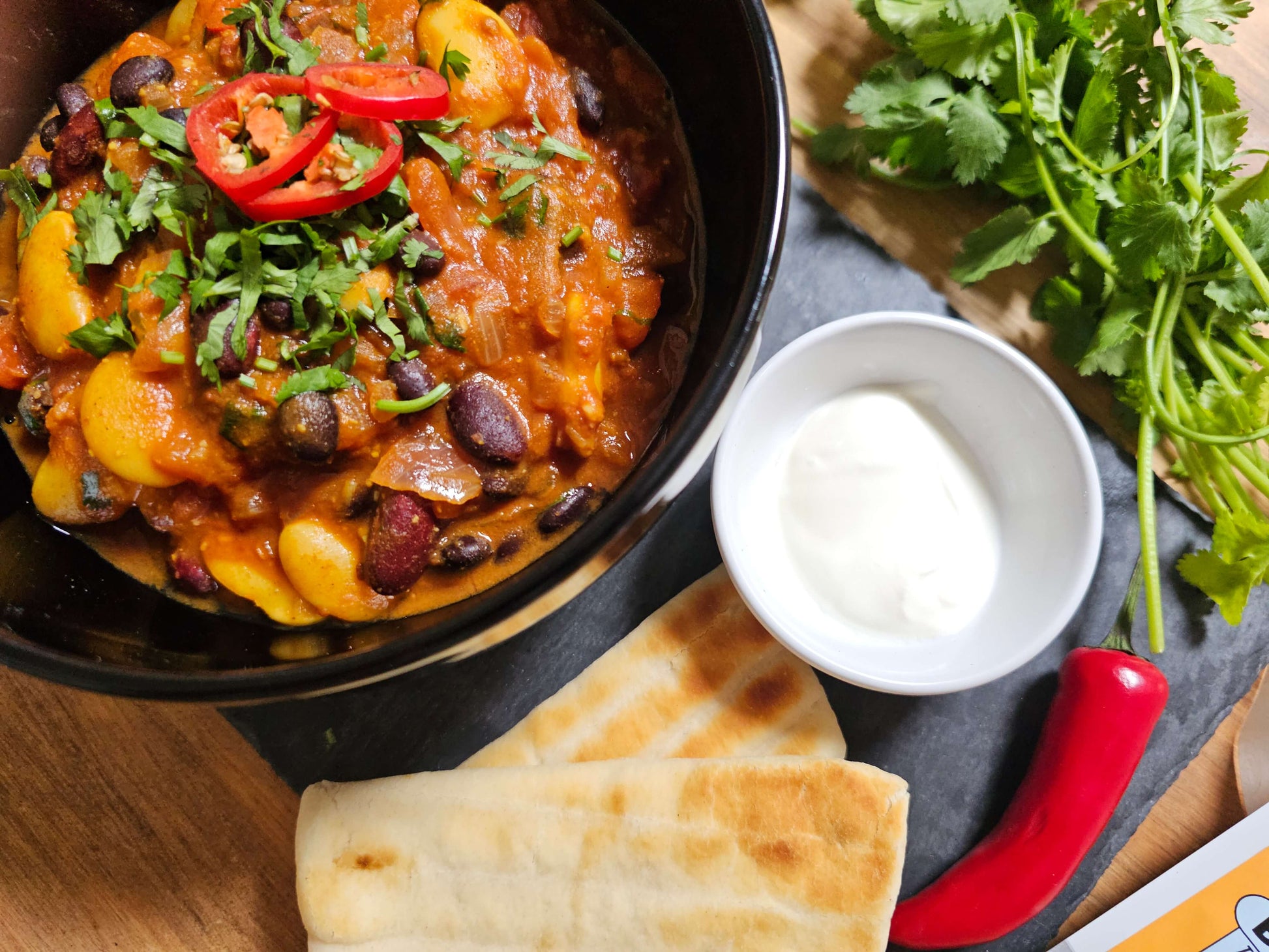 Three bean curry in a black bowl. Next to is is some flatbread, sour cream, fresh chilies and coriander