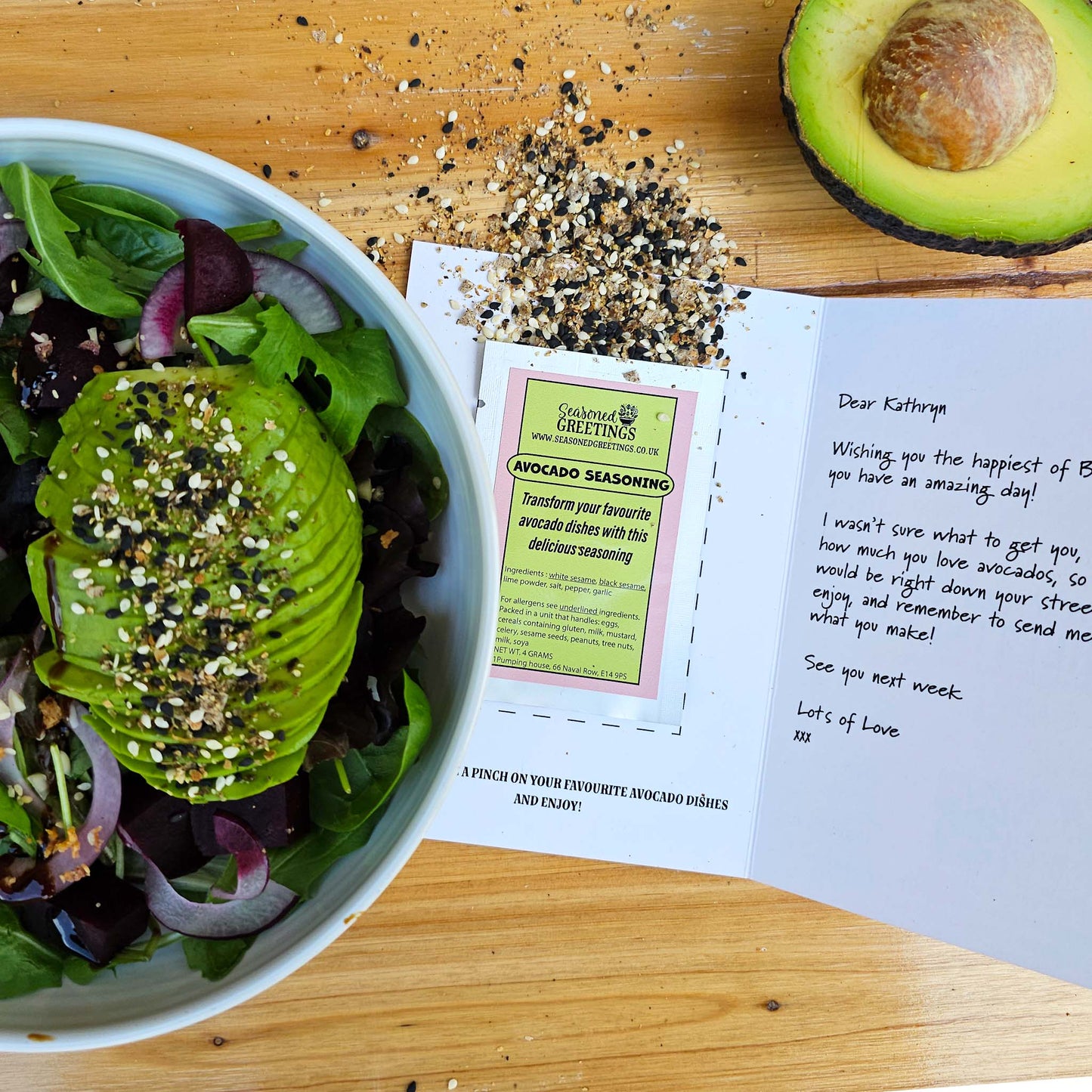 an avocado salad in a white bowl. Next to it is an opened seasoned greetings card showing avocado seasoning spilling out of the sachet on to the wood table. Above it is half an avocado